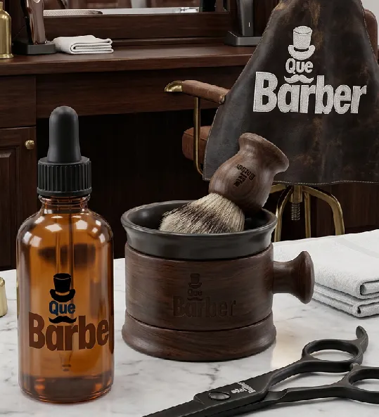 Productos de Que Barber en la barbería: un frasco de aceite, la taza y la brocha de afeitar con el logo, unas tijeras y la capa bordada al fondo.
